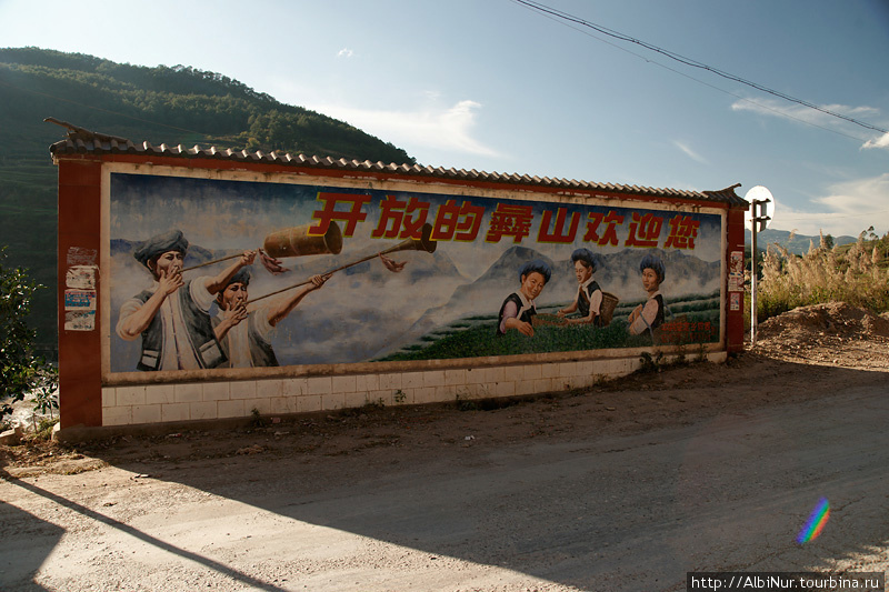 Проезжаем какую-то национальную деревню, на всех плакатах один мотив — мужчины дудят в дудки и пляшут, женщины работают. Джинхонг, Китай